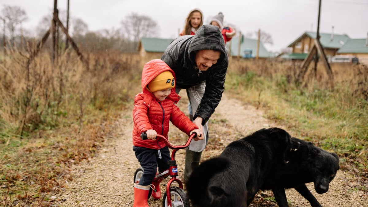 Dad helps son ride a bike on a path with family and dog, capturing a joyful outdoor moment.