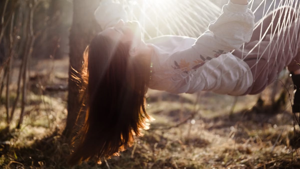 Young woman with long hair lies playfully in a sunlit hammock outdoors, embracing nature and sunlight.